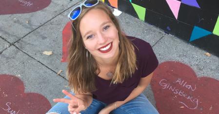 La fille au pair est assise en tailleur par terre, regarde vers le haut et fait le signe de la paix avec sa main.